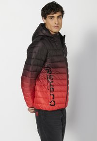 Giacca imbottita con gradiente dal nero al rosso, cappuccio, chiusura con zip, dettaglio del logo sul lato e texture liscia. Indossata con jeans scuri.