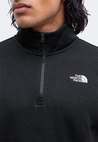 Gros plan d'une personne portant un pull noir à fermeture éclair au niveau de la poitrine avec le logo The North Face sur la poitrine.