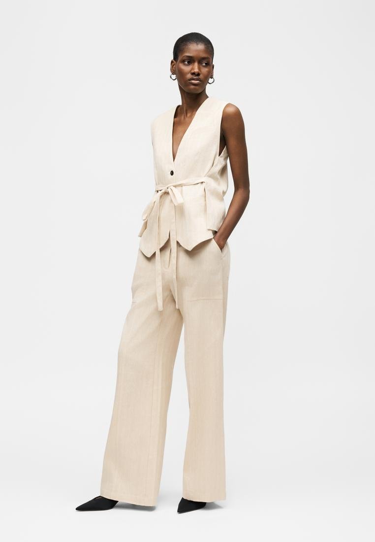 Femme posant en gilet beige sans manches avec ceinture et pantalon large assorti, portant des chaussures noires à bouts pointus et des boucles d'oreilles argentées en forme de créoles.