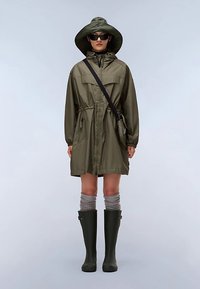 Giacca impermeabile verde oliva con coulisse in vita, grandi tasche e cappuccio a tesa larga, indossata sopra calzini grigi al ginocchio e stivali da pioggia verdi.