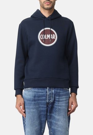 Mann trägt einen marineblauen Hoodie mit „COLMAR“-Logo auf der Brust und blaue ausgewaschene Jeans vor weißem Hintergrund.