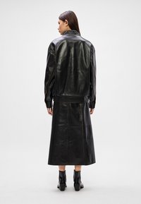 Crna kožna jakna s malo prevelikim krojem, koja ima vidljivo šivanje i visoki ovratnik, u kombinaciji s odgovarajućom crnom kožnom suknjom.