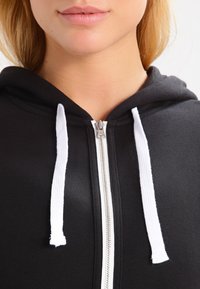 Sort hættetrøje med lynlås og hvide snore, der har en glat tekstur og en metal lynlåslukning foran.