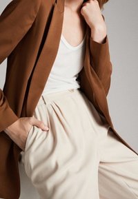 Brązowa dopasowana marynarka na biały top na ramiączkach, w połączeniu z beżowymi plisowanymi spodniami. Tkaniny wyglądają na gładkie o wyrafinowanym wykończeniu.