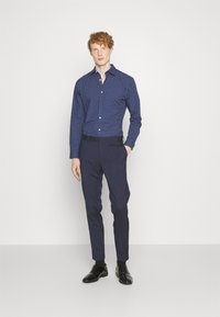Camicia button-up blu navy con pois bianchi, slim fit, abbinata a pantaloni scuri e scarpe nere lucide, in piedi su uno sfondo chiaro.