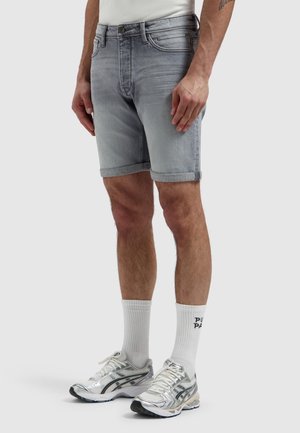 Man met lichtgrijze denimshorts, witte sportsokken met zwarte tekst, zilveren hardloopschoenen en een wit shirt.