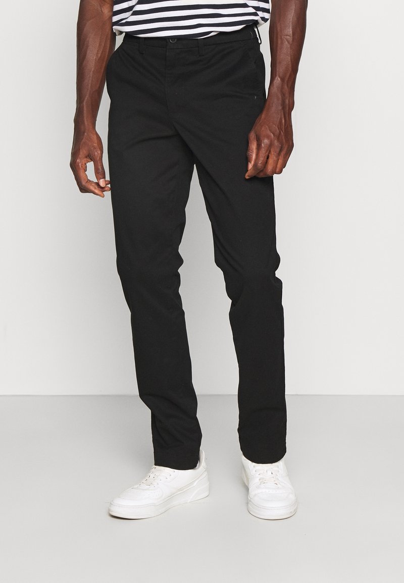 Pantalon en coton noir à coupe droite, associé à des baskets blanches. La tenue comprend une chemise rayée et une silhouette simple.