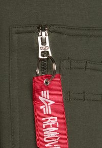 Indumento verde oliva con una cerniera e un tirante argentato etichettato "ALPHA." Include un'etichetta di tessuto rossa con testo bianco e un portachiavi.