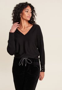 Pull noir à col en V avec des manches longues, présentant un léger scintillement et des accents de boutons aux poignets. Associé à un pantalon en velours côtelé noir avec cordon de serrage.