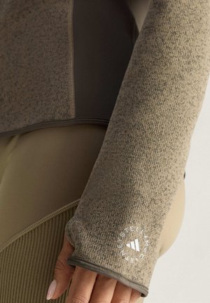 Dlouhý rukáv v béžové a hnědé barvě, s texturovanou pletenou látkou a logem na rukávu. Design zahrnuje kontrastní panely.