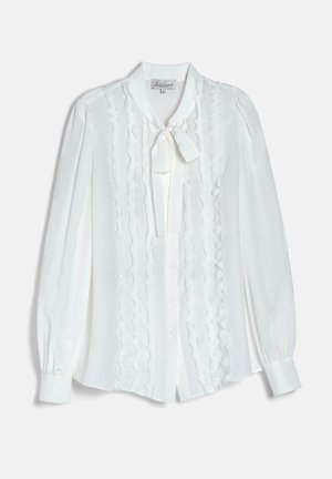 Kvinde med kort brunt hår bærer en hvid bluse med flæser og sløjfe, sorte jeans, store guldringe og sort bælte med firkantet juvelspænde.