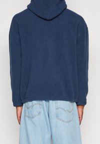 Felpa con cappuccio in pile blu navy dal tessuto morbido, vestibilità comoda e tasca a marsupio. Abbinata a pantaloni di denim azzurro chiaro.