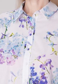 Chemise à boutons florale en tissu léger, ornée de fleurs bleues et violettes sur fond blanc. Col pointu et boutons visibles.