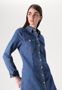 Vestido em denim azul médio com mangas longas, frente abotoada, dois bolsos no peito e detalhes em costura dourada. Ajuste estruturado.