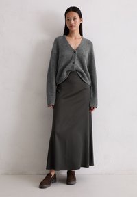 Grijze gebreide cardigan met knopen, gecombineerd met een donkergroene lange rok en brun suede schoenen. Glad texturen in het hele outfit.