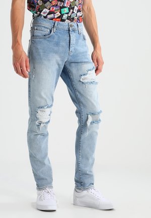 Lichtblauwe jeans met een verweerde look en gescheurde kniedetails, standaard ontwerp met vijf zakken en een ontspannen pasvorm. Gecombineerd met witte sneakers.