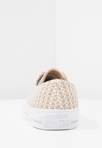 Zapatilla de lona texturizada beige con un patrón tejido, suela de goma blanca y un logo en el talón. Diseño tipo slip-on con un perfil bajo.