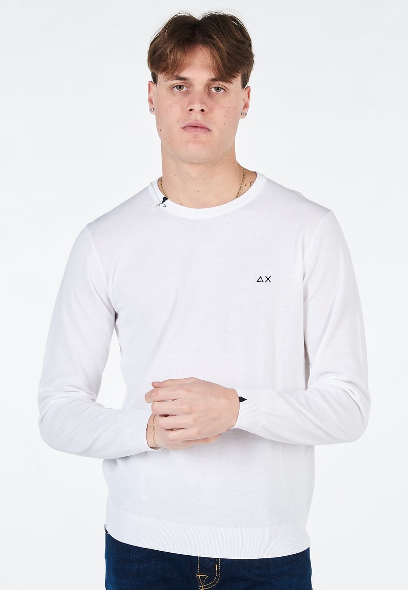 Maglione a maniche lunghe bianco in maglia con scollatura tonda, logo nero sul lato sinistro del petto e polsini e orlo a coste, abbinato a jeans in denim blu.