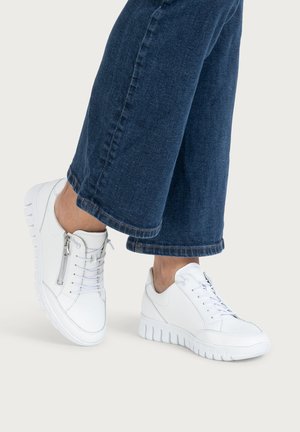 Person wearing white lace-up sneakers with side zipper details and cropped dark blue denim jeans against a plain background.