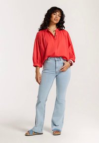 Crvena lagana bluza s naduvanim rukavima, uparena s lagano plavim trapezastim trapericama sa bočnim prorezima. Model nosi sandale s remenčićem.