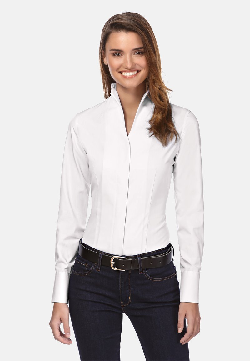 Chemise blanche à manches longues avec un col haut, un design ajusté et des détails de couture verticaux, associée à un jean foncé et une ceinture noire.