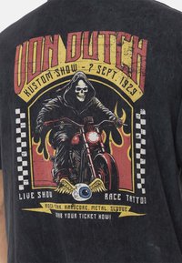 T-shirt noir présentant un graphique d'un cavalier squelettique sur une moto, entouré de flammes, avec un texte en rouge et jaune audacieux, et des accents en damier.