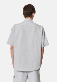 Camicia a maniche corte in tessuto grigio chiaro con vestibilità rilassata, colletto a punta e cuciture rinforzate lungo le cuciture delle spalle.