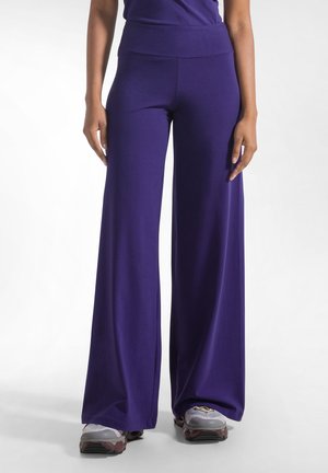 Vrouw die paarse broek met hoge taille en brede pijpen draagt met sneakers, staand tegen een effen witte achtergrond.