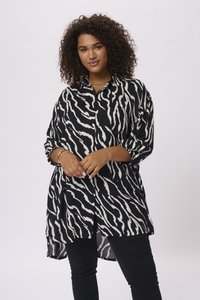 Chemise à rayures zèbres noir et blanc, à manches longues et retroussées ; tissu léger ; ourlet arrondi ; associée à un pantalon noir.