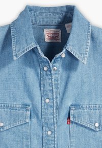 Vaaleansininen farkkupaita, jossa on klassinen kaulus, nepparit, kaksi rintataskua ja pieni punainen logolappu vasemmassa taskussa.