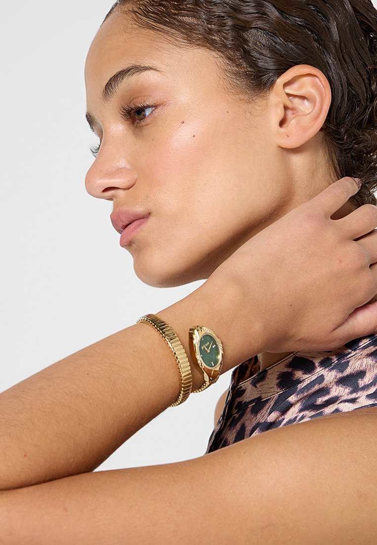 Gouden horloge met een groene wijzerplaat, ronde kast en dunne wijzers; vergezeld door een gouden armband met textuur. Fijn ontwerp met contrasterende elementen.