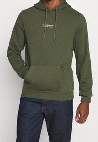 Sudadera verde hecha de una mezcla de algodón. Presenta un bolsillo frontal, cordones en la capucha y texto impreso en el pecho. Textura suave.