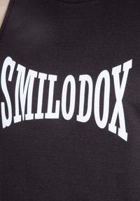 Haut sans manches noir avec un texte incurvé et audacieux en blanc affichant "SMILODOX", fabriqué dans un matériau doux et extensible.