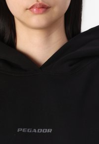 Hanorac negru cu glugă, având textul "PEGADOR" scris cu alb. Materialul este neted, cu o vedere detaliată a gâtului și a glugii.
