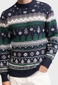 Marineblauer Strickpullover mit grünen Bäumen, weißen und grauen Schneeflocken, geometrischen Mustern, getragen von einer Person, die eine Hand in der Tasche der weißen Hose hat.