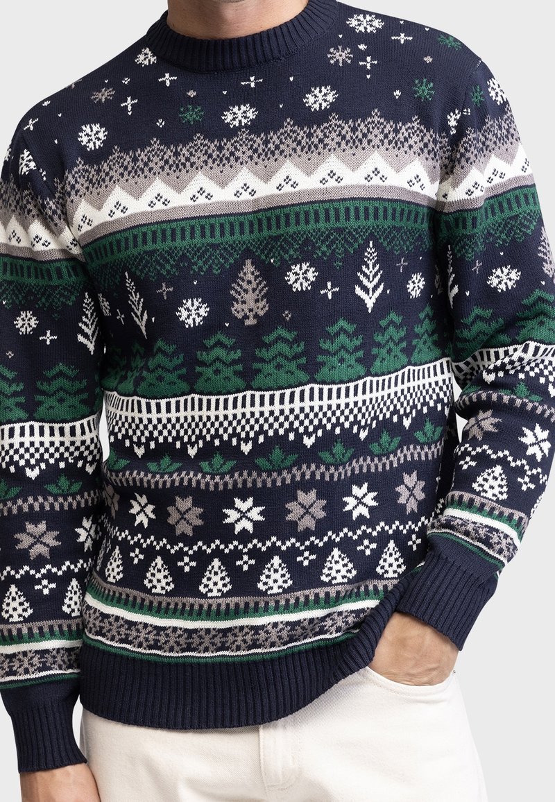Marineblauer Strickpullover mit grünen Bäumen, weißen und grauen Schneeflocken, geometrischen Mustern, getragen von einer Person, die eine Hand in der Tasche der weißen Hose hat.