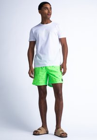 Biała bawełniana koszulka, neonowo zielone szorty kąpielowe w czarne wzory oraz brązowe klapki. Model stoi na jasnym tle.