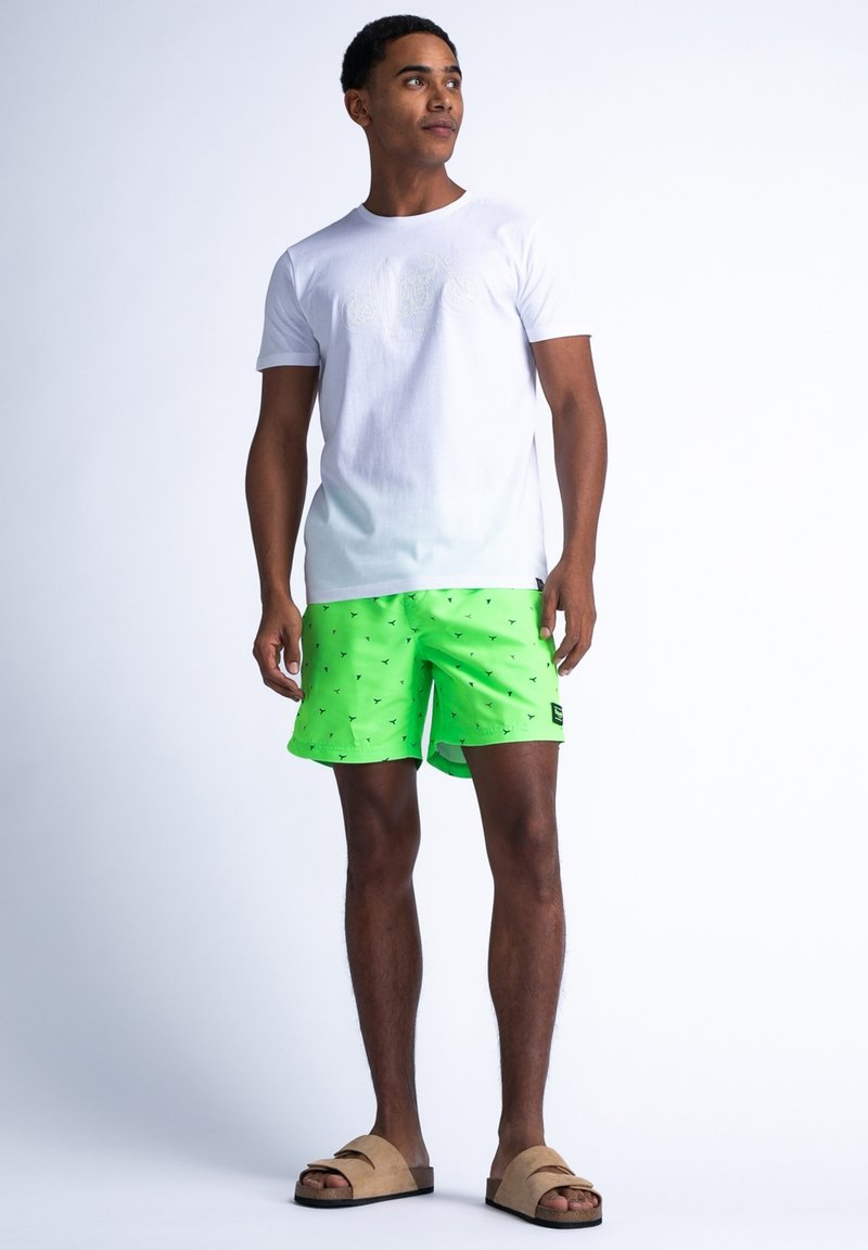 Biała bawełniana koszulka, neonowo zielone szorty kąpielowe w czarne wzory oraz brązowe klapki. Model stoi na jasnym tle.