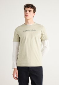 Světle zelené bavlněné tričko s šedým logem "calvin klein", vrstvené přes bílé triko s dlouhým rukávem, doplněné tmavými kalhotami.