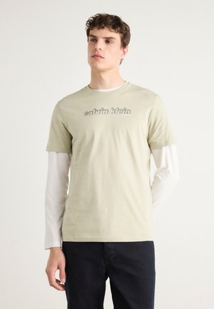 Hele roheline puuvillane t-särk, millel on hall "calvin klein" logo, kihiti valge pika varrukaga särgi kohal, võrreldes tumedate pükstega.