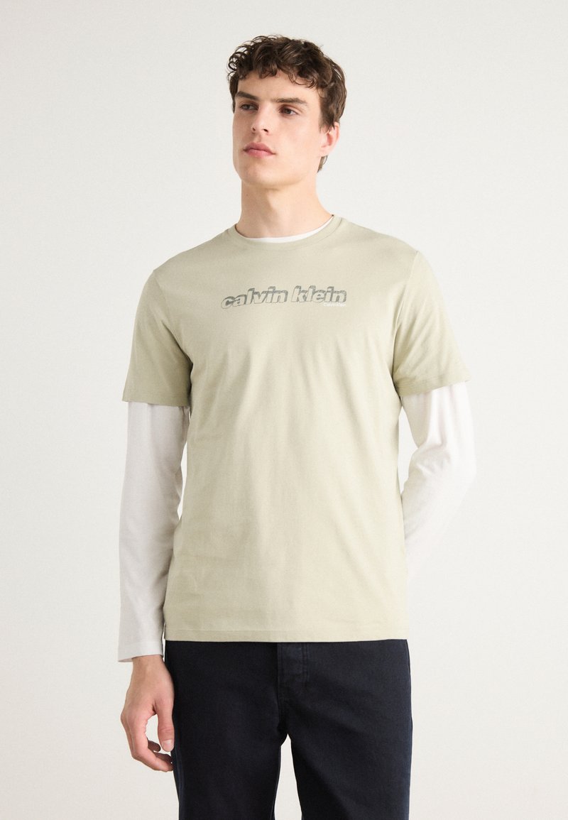 Světle zelené bavlněné tričko s šedým logem "calvin klein", vrstvené přes bílé triko s dlouhým rukávem, doplněné tmavými kalhotami.