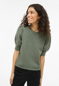 Jonge vrouw met krullend haar die glimlacht, draagt een olijfgroene trui met korte mouwen en een zwarte broek, kijkt naar links.