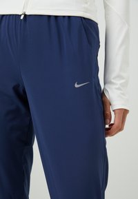 Granatowe spodnie sportowe o gładkiej fakturze, z elastycznym pasem oraz odblaskowym szarym logo Nike na lewej udzie.