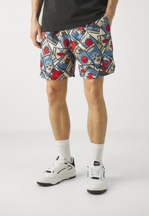 Homme portant un short graphique coloré avec des motifs de basketball et de style bande dessinée, chaussettes blanches et baskets blanches Puma sur un fond uni.