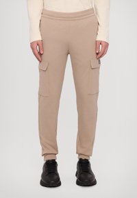 Beige cargo joggere med elastisk midje, to sidelommer og smale mansjetter, laget av et mykt materiale. Svarte joggesko fullfører antrekket.