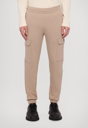Beige cargo joggers with elastic waistband, two side pockets, and tapered cuffs, made from a soft fabric. Black sneakers complete the look.