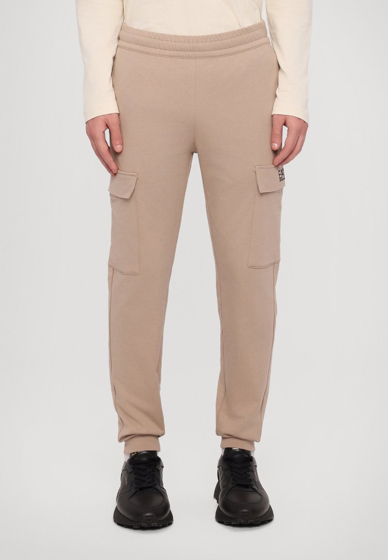 Beige cargo joggere med elastisk midje, to sidelommer og smale mansjetter, laget av et mykt materiale. Svarte joggesko fullfører antrekket.
