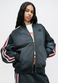 Γυναίκα με σκούφο μπουφάν με φερμουάρ και αντίστοιχα παντελόνια με κόκκινες και λευκές ρίγες, με τα χέρια στις τσέπες, κοιτάζει με αυτοπεποίθηση μπροστά.