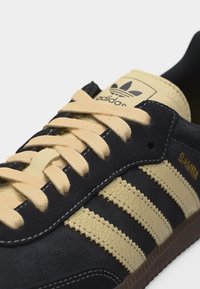 Κοντινό πλάνο μαύρου αθλητικού παπουτσιού Adidas Samba με μπεζ κορδόνια, τρεις μπεζ ρίγες, σόλα gum και λογότυπο Adidas στη γλώσσα.