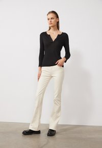 Haut noir à manches longues avec un col en V, porté avec un pantalon évasé blanc cassé et des chaussures noires. Texture lisse, design cintré et détails subtils.